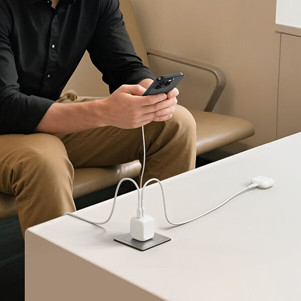 A person sitting on a bench using a smartphone while it charges from a white wall charger plugged into a table outlet, with another device cable resting on the tabletop.