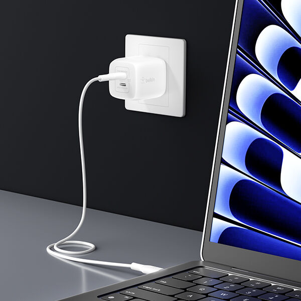 A white wall charger plugged into an outlet, with a cable running to a laptop on a desk, set against a dark wall background.