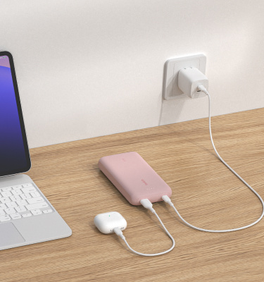 A pink Belkin BoostCharge Power Bank 20K with Integrated Cable is placed on a wooden surface, charging a pair of AirPods while being recharged from a wall outlet, with a tablet nearby.