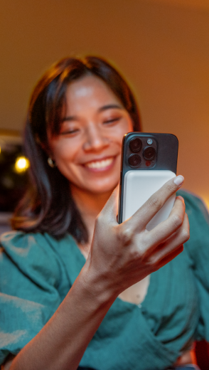 A smiling woman holds an iPhone with a Belkin BoostCharge Pro Magnetic Power Bank with Qi2 15W attached, showcasing the ease of portable charging, in a warmly lit setting.
