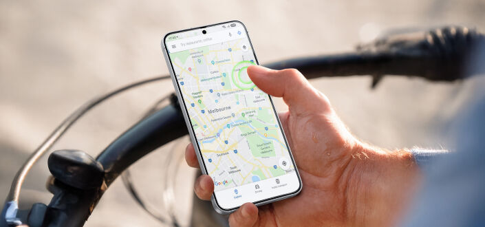 Person holding a smartphone while riding a bicycle, with a navigation map open on the screen and the bike&rsquo;s handlebars visible in the background.&rdquo;