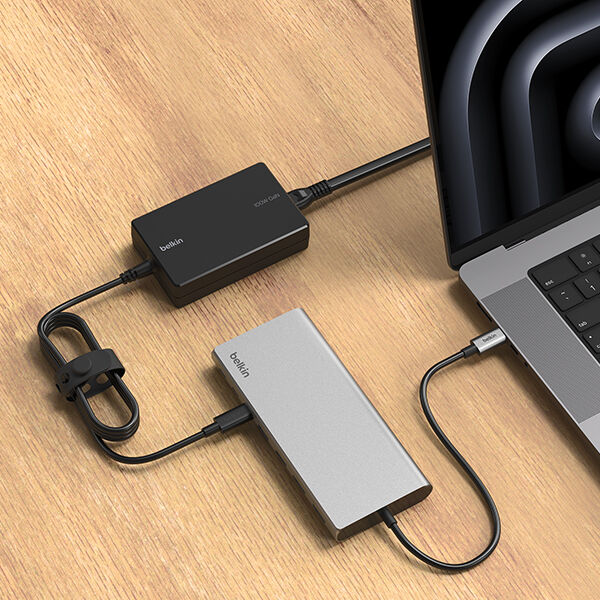 A close-up of a modern workspace featuring a MacBook connected to a Belkin docking station and a power adapter. The docking station and power adapter are neatly arranged on a wooden desk, with cables organized for a clean and efficient setup.