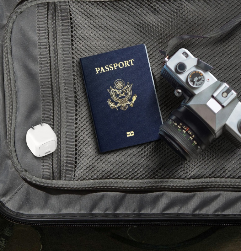 The image shows the Belkin BoostCharge Compact USB-C Wall Charger 30W with foldable prongs placed in a suitcase alongside a passport and a camera, highlighting its compact, travel-friendly design.