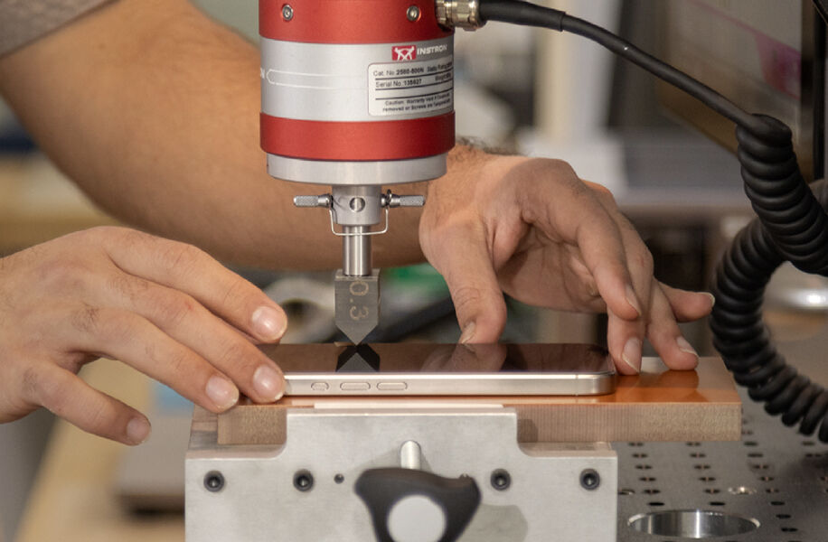 Close-up of a smartphone secured on a testing platform while a mechanical press applies force to the screen, demonstrating laboratory durability and impact testing.