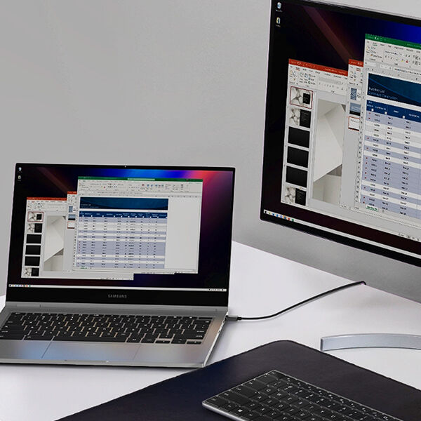 A Samsung laptop connected to an external monitor via a single cable, displaying an extended screen setup with Excel and PowerPoint applications open. A wireless keyboard is placed on a black desk mat, creating a clean and organized workspace.