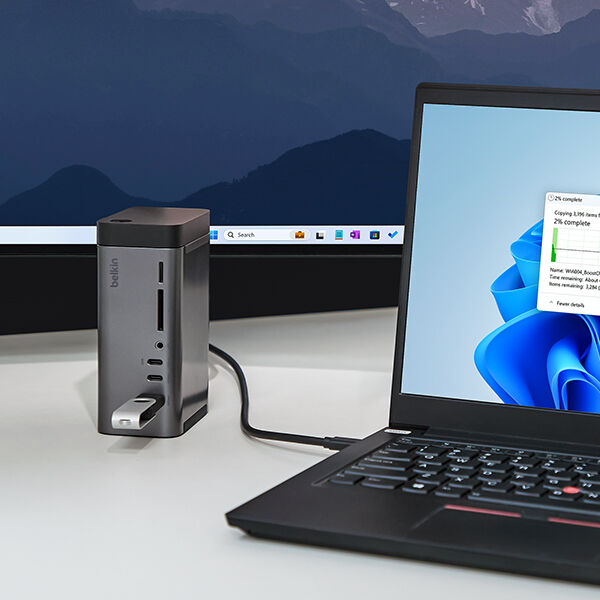 A workspace setup featuring a Belkin docking station connected to a laptop running Windows 11, with a file transfer progress window displayed on the screen. The docking station has multiple ports, including USB and SD card slots, with a USB flash drive inserted. A large external monitor is visible in the background, displaying a Windows desktop. The setting is clean and modern, emphasizing connectivity and efficiency.