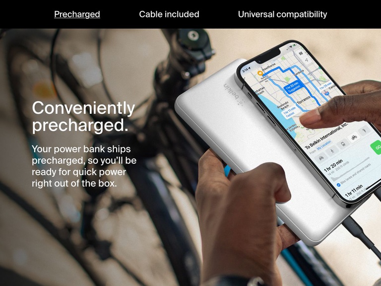 A person charges their smartphone using a white Belkin BoostCharge Power Bank 20K while navigating on a bike, highlighting its convenient precharged feature for immediate use.
