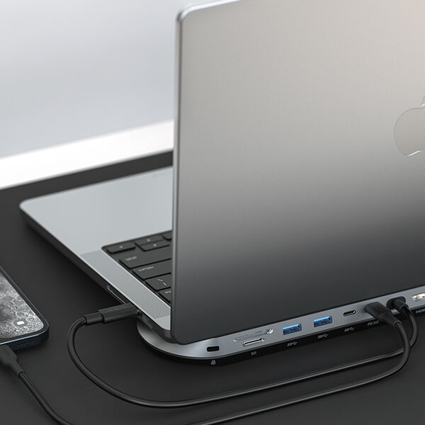 A silver MacBook connected to a sleek, curved docking station with multiple ports, including USB-A, USB-C, and SD card slots. The docking station is placed on a black desk, with a smartphone partially visible to the side, highlighting a clean and efficient workspace setup.