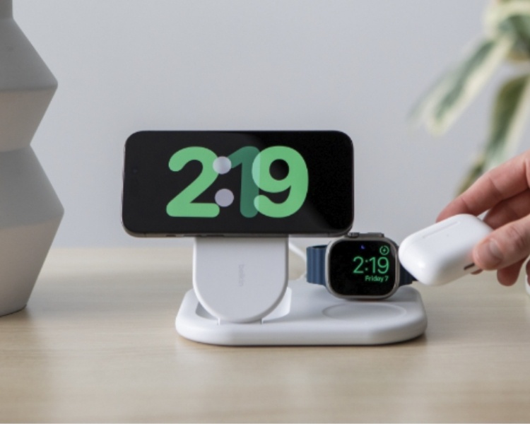 Eine Person platziert einen kabellosen Kopfhörer auf einer Belkin 3-in-1-MagSafe-Ladestation, auf der eine Smartwatch und ein Smartphone im Querformat aufgeladen werden. Das Ladegerät befindet sich auf einer Holzoberfläche mit einer Pflanze und anderer Dekoration im Hintergrund.