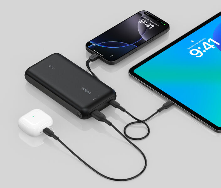 A black Belkin BoostCharge Power Bank 20K with Integrated Cable is shown charging an iPhone, an iPad, and AirPods simultaneously on a reflective surface.