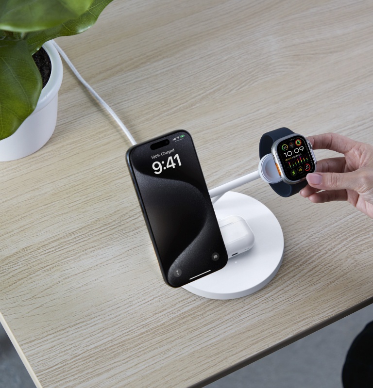 A Belkin BoostCharge Pro 3-in-1 Wireless Charging Stand with Qi2 15W is shown on a wooden desk, charging an iPhone, AirPods, and an Apple Watch being placed on the stand by a hand.