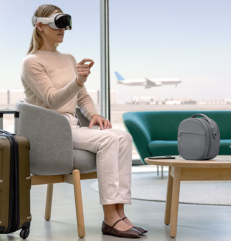 A woman wearing Apple Vision Pro with the travel bag for Apple Vision Pro besides her at an airport. 