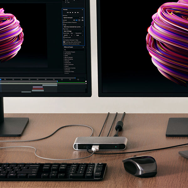 A dual-monitor workstation setup with a Belkin docking station connecting peripherals, including a keyboard and mouse. The screens display a digital art project featuring a vibrant 3D-rendered object. Cables are neatly arranged, emphasizing an organized and efficient workspace for creative professionals.