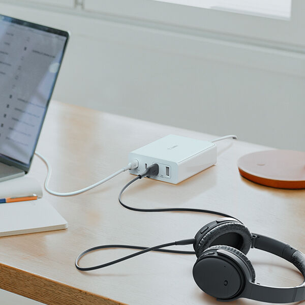 A minimalist workspace featuring a wooden desk with a laptop, a white Belkin charging hub connected to multiple devices, and a pair of black over-ear headphones. Natural light from a nearby window enhances the clean and organized setting.