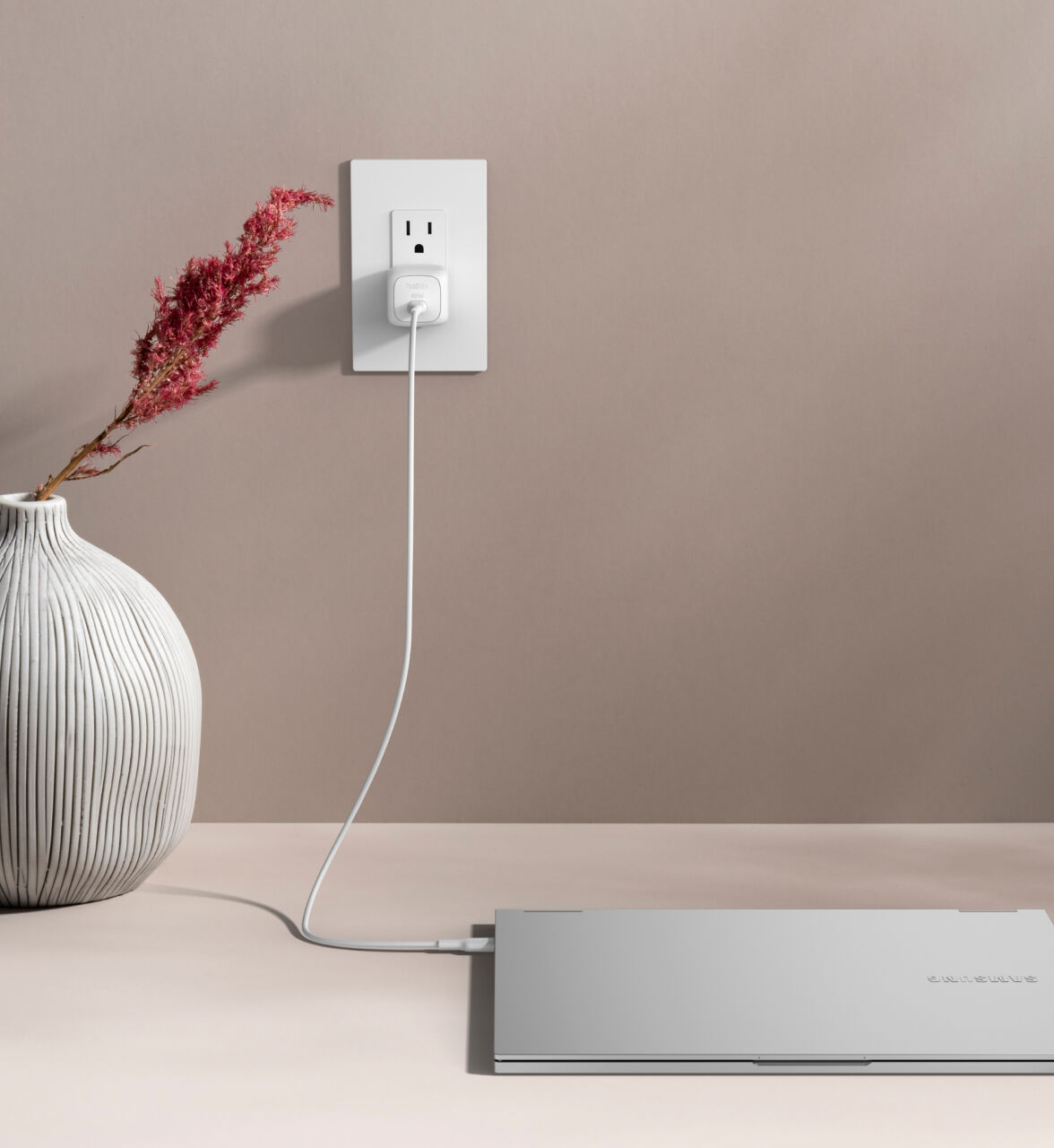 A Belkin BoostCharge Compact USB-C Charger 45W is plugged into a white wall outlet, charging a closed silver laptop on a beige surface with a white cable, next to a decorative vase with dried flowers.