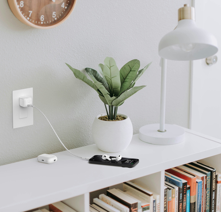 L'image montre le chargeur mural USB-C BoostCharge Compact 30 W de Belkin branché sur une prise murale, chargeant rapidement un iPhone sur une table voisine avec des AirPods placés sur le dessus, à côté d'une petite plante en pot.