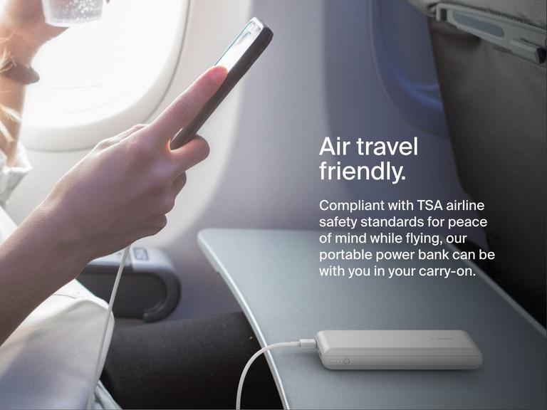 A person uses a white Belkin BoostCharge Power Bank 20K to charge their phone on an airplane tray table, highlighting its TSA-compliant, travel-friendly design.