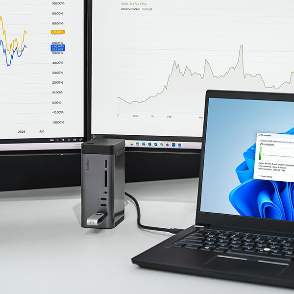 A Belkin docking station connected to a laptop and USB drive, with dual monitors displaying financial graphs and charts in the background.