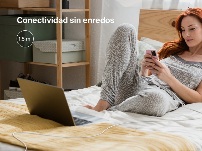 Una mujer se relaja echada en la cama usando su iPhone conectado a un cable Belkin BoostCharge USB-C con conector Lightning, mostrando los 1,8 m de longitud sin enredos, ideal para un mayor alcance y una carga flexible.