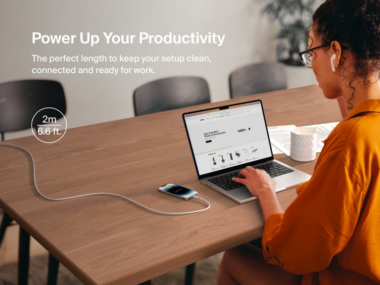 A woman works at a desk with her iPhone charging via a 2-meter (6.6 ft) Belkin BoostCharge Braided USB-A Cable with Lightning Connector, ideal for maintaining a clean, connected workspace.