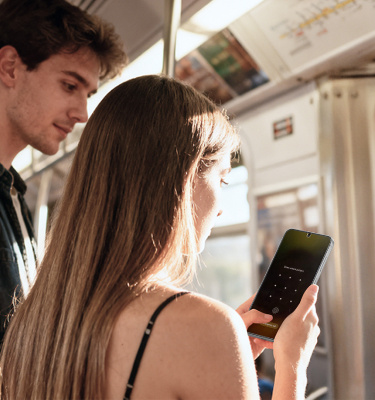 Woman standing on a train holding and looking at a smartphone, while another passenger stands behind her, with sunlight streaming through the window.