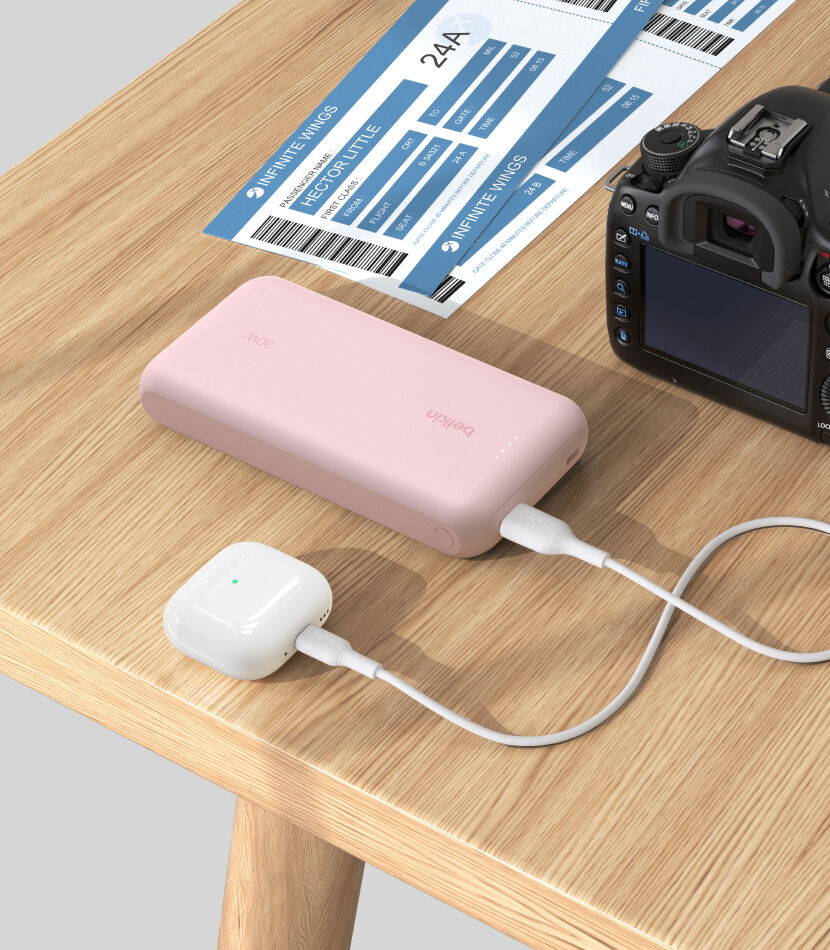 A pink Belkin BoostCharge Power Bank 20K with Integrated Cable is placed on a wooden table, charging a pair of white AirPods, with a DSLR camera and boarding passes nearby. A pink Belkin BoostCharge Power Bank 20K with Integrated Cable is placed on a wooden table, charging a pair of white AirPods, with a DSLR camera and boarding passes nearby.