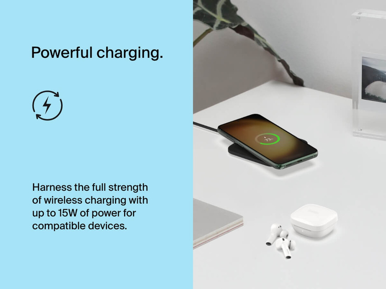 A smartphone charges on the Belkin BoostCharge Universal Easy Align Wireless Charging Pad 15W on a white desk, demonstrating 15W wireless power alongside nearby earbuds and workspace accessories.