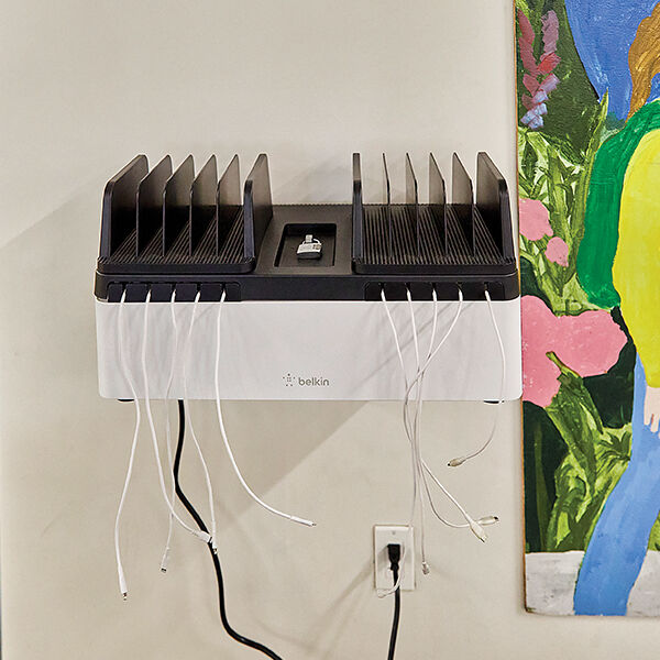 A wall-mounted Belkin charging station with multiple vertical slots and several white charging cables hanging from the front. The unit is placed next to a colorful mural featuring flowers and a partially visible person.