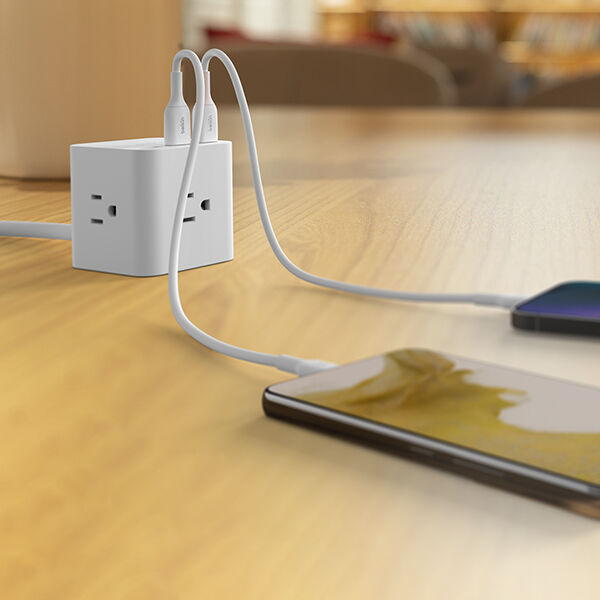 A cube-shaped power outlet on a wooden table with two USB cables plugged in, charging two smartphones placed on the tabletop.