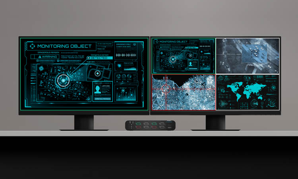 Dual-monitor setup displaying surveillance and monitoring interfaces, including maps, satellite imagery, and data dashboards. A Belkin multiclave KVM switch is positioned between the monitors, enabling secure control and source switching across multiple network environments.