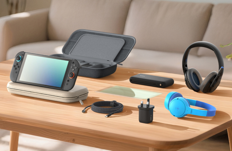 A variety of Nintendo Switch 2 accessories are neatly arranged on a wooden table in a cozy living room setting. The items include a Nintendo Switch 2 console resting on a carrying case, an open protective storage case, a power bank, a screen protector, a USB-C charging cable, a wall charger, and two pairs of headphones (one black and one blue). Soft natural light streams in through the window, creating a warm and inviting workspace for gaming.