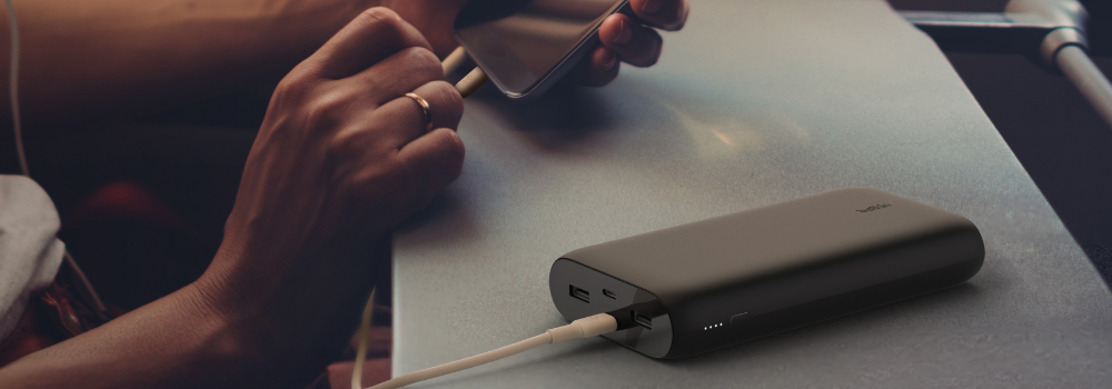 A person charging their smartphone using a black Belkin power bank on an airplane tray table. The power bank has multiple ports and LED indicators, highlighting its portability and convenience for travel. The background shows a dimly lit cabin, creating a comfortable in-flight charging setup.