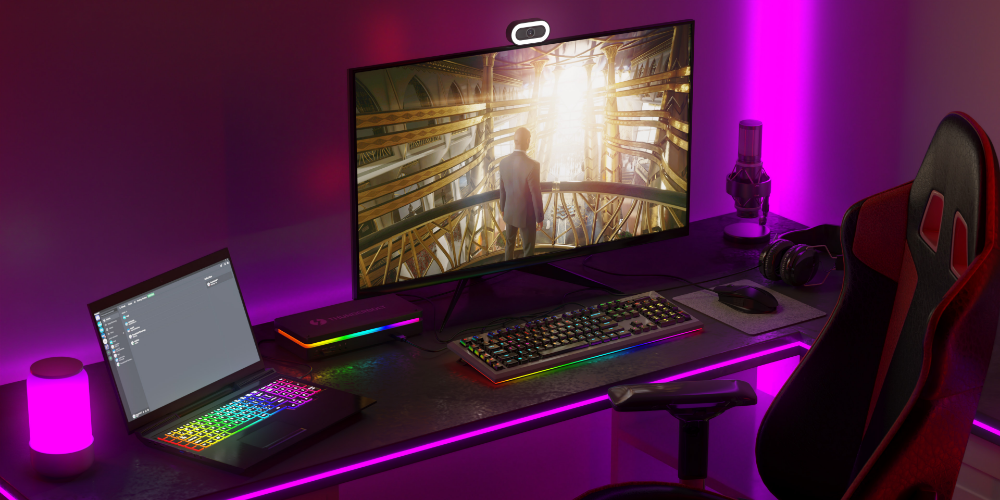 A futuristic gaming and streaming setup featuring a dark desk illuminated by vibrant purple LED lighting. The setup includes a large monitor displaying a sci-fi scene, a gaming laptop with an RGB keyboard, a mechanical keyboard, and a mouse with customizable lighting. A webcam is mounted on top of the monitor. Additional peripherals include a microphone, a pair of headphones, and a sleek external storage device. A gaming chair with red and black accents complements the setup, creating an immersive, high-tech atmosphere.