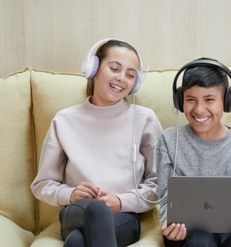 Two kids both wearing the headphones and the two headphones are connected with each other. They are listening to music while using an iPad.