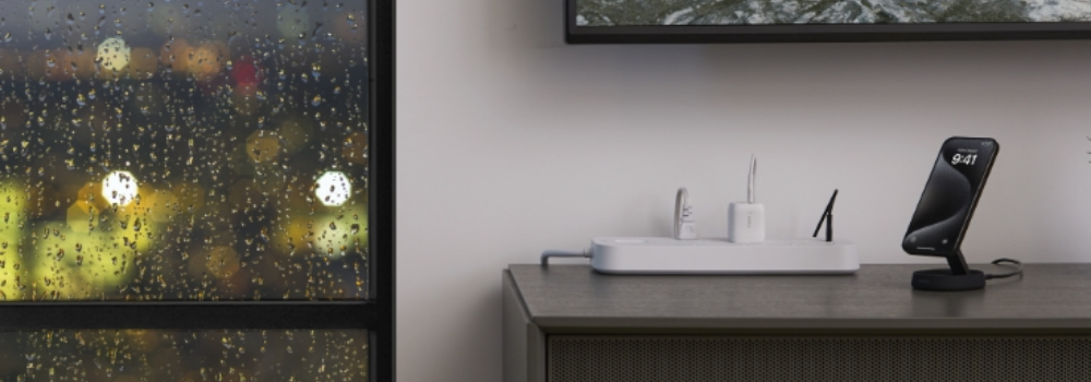 A modern power setup on a cabinet featuring a Belkin power strip charging multiple devices, with a smartphone propped on a wireless charging stand beside a window with rain droplets at night.