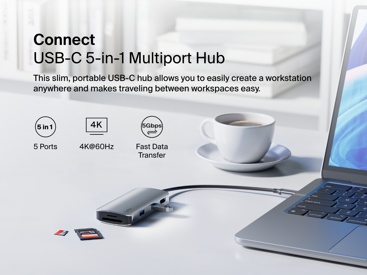 A sleek, silver Belkin Connect USB-C 5-in-1 Multiport Hub is connected to a laptop on a white desk, with SD cards nearby and a cup of coffee in the background, highlighting its portability and functionality.
