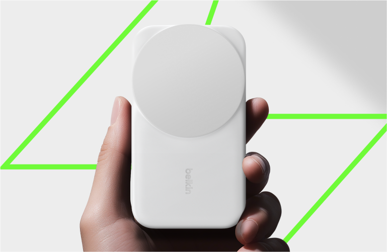Una mano tiene una power bank wireless magnetica Belkin di colore bianco, su sfondo luminoso con contorno geometrico verde.