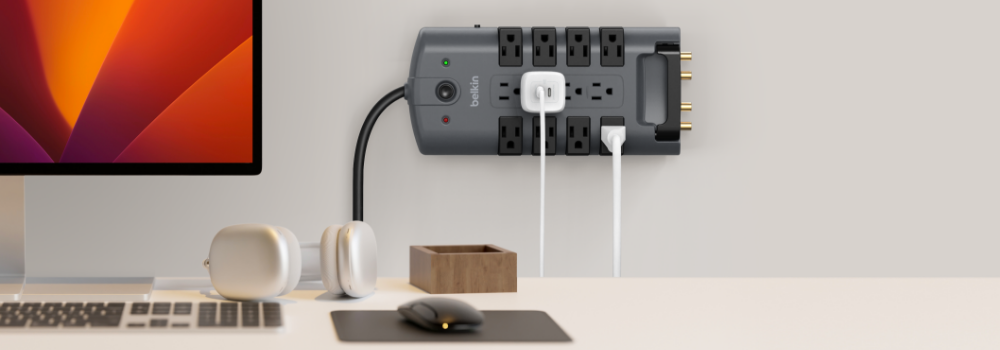 A Belkin surge protector mounted on a wall above a minimalist desk setup, powering multiple devices including a desktop computer, with headphones and accessories neatly arranged nearby.