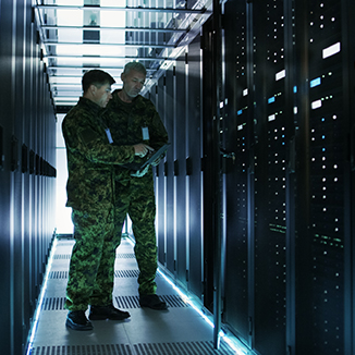 Two military personnel in camouflage uniforms examine a tablet while standing in a high-security data center server room. Rows of illuminated server racks line both sides.