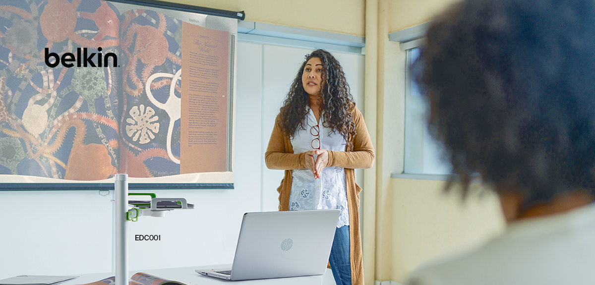 A teacher presenting in a modern classroom using Belkin's EDC001 presentation tool. The teacher is standing in front of a projected image of a colorful educational diagram, with a laptop on the desk. The Belkin device is visible in the foreground, showcasing its functionality in enhancing classroom presentations.
