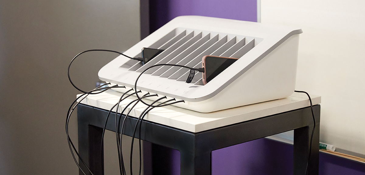A white multi-device charging station with vertical slots, holding several smartphones connected with charging cables, placed on a small table against a purple and white wall.