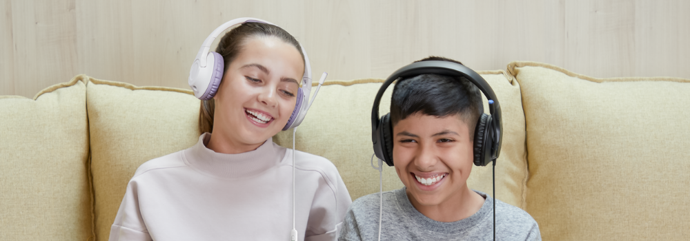 Two children sitting on a beige couch, smiling and wearing Belkin headsets with microphones. The girl on the left is wearing a light pink top and lavender headset, while the boy on the right is wearing a gray shirt and black headset. They appear to be enjoying a shared activity, possibly gaming or learning online.
