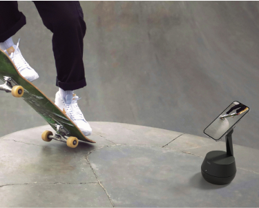 This image captures a skateboarder performing a trick in a skatepark while being recorded by a smartphone mounted on a sleek, black motion-tracking stand. The stand is positioned to follow the movement dynamically, emphasizing its ability to track and record action seamlessly. The background features a textured skatepark surface, highlighting an active and adventurous setting.