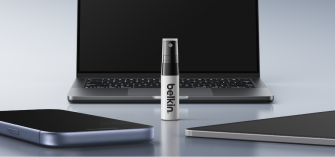 Belkin screen protection spray bottle standing on a desk between a laptop and two smartphones, highlighting device care and cleaning.
