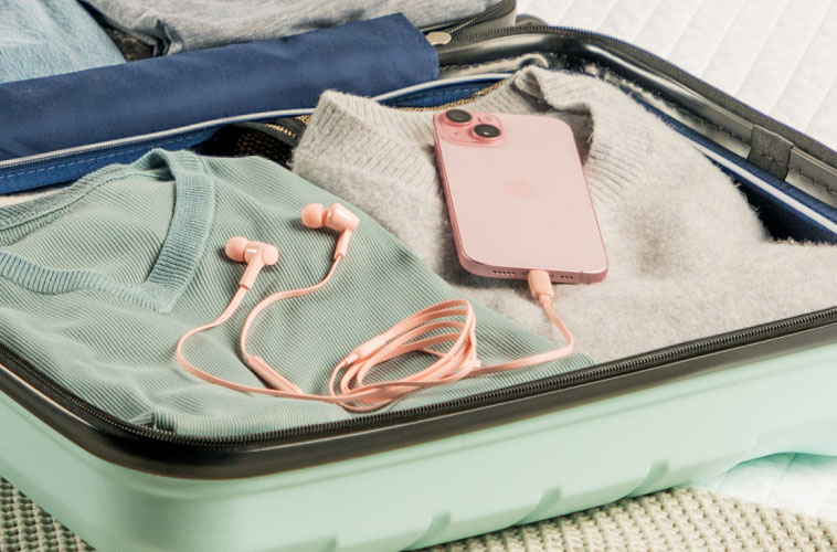 Pink wired earphones connected to a matching pink smartphone, resting on neatly folded clothes inside an open mint green suitcase.