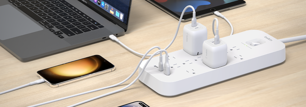 A white Belkin surge protector on a wooden desk powering multiple devices, including a laptop, smartphone, and tablet, with several USB-C and wall chargers plugged in.