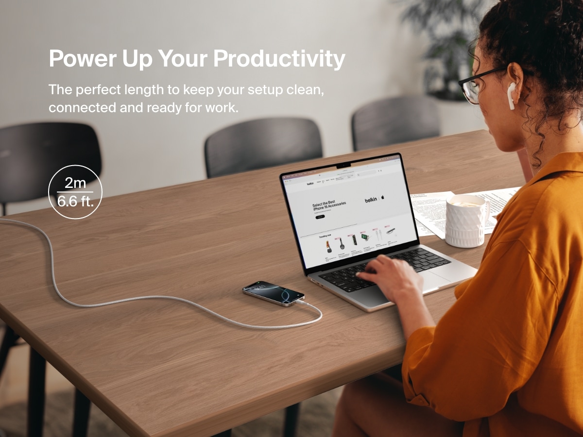 A woman works at a desk while her phone charges with a 2-meter (6.6 ft) Belkin BoostCharge Pro Braided USB-A to USB-C Cable 15W, offering extended reach and a clutter-free workspace.