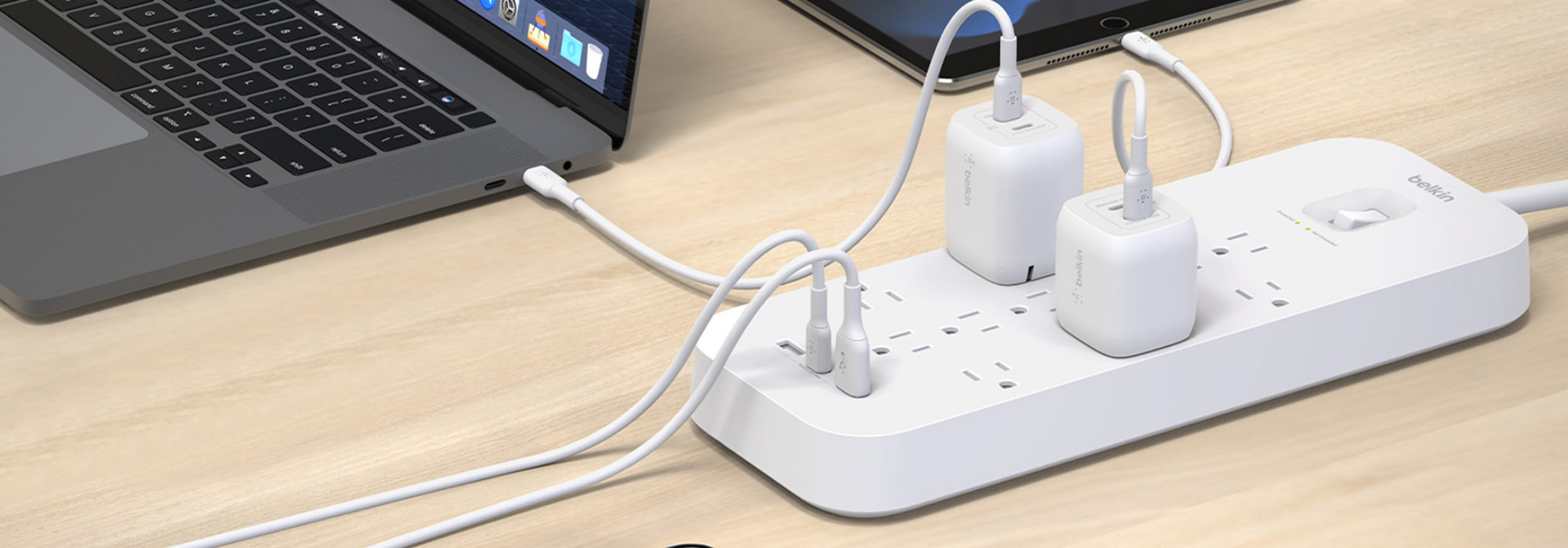 A white Belkin surge protector power strip on a wooden surface, with multiple devices plugged in, including two Belkin wall chargers. The power strip is connected to a MacBook and a tablet via USB-C cables. The power strip features multiple outlets and a power switch with an indicator light.