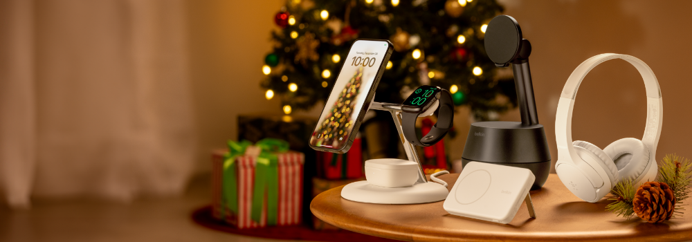 Holiday scene with Belkin tech gifts displayed on a table, including wireless charging stands holding a phone and smartwatch, a charging pad, and over-ear headphones, with a decorated Christmas tree and wrapped presents in the background.