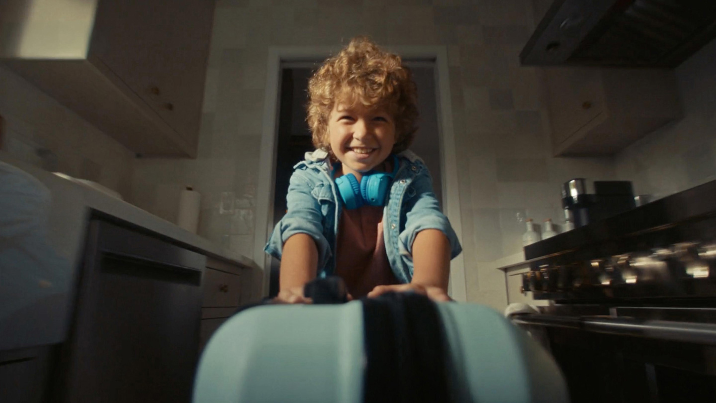 Young boy with curly hair smiling and pushing a blue suitcase toward the camera in a kitchen, wearing a denim jacket and bright blue headphones around his neck.
